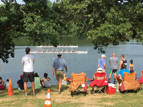 A group of people standing and seated watching two white 8 boats racing down Noxontown Pond.