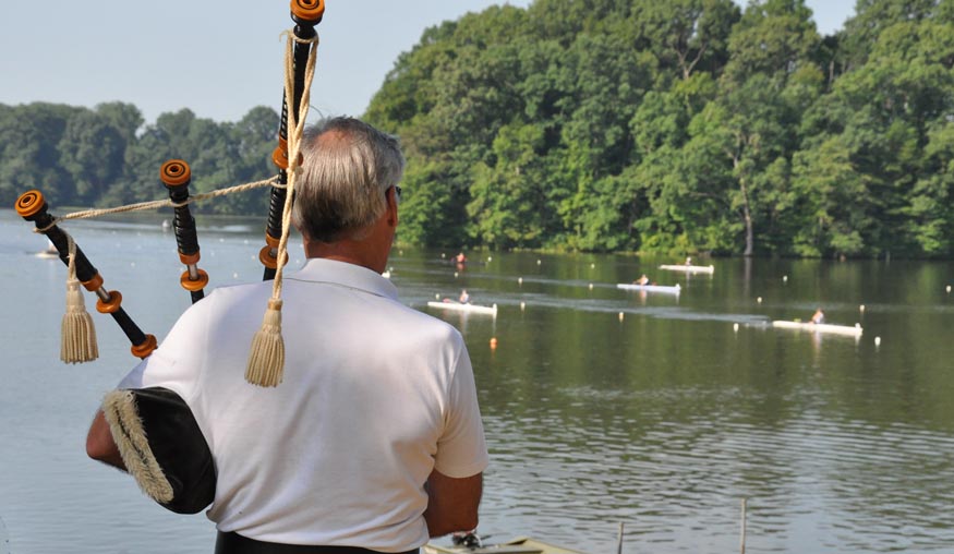 A man playing bagpipes overlooking a boat where several singles are rowing a race.